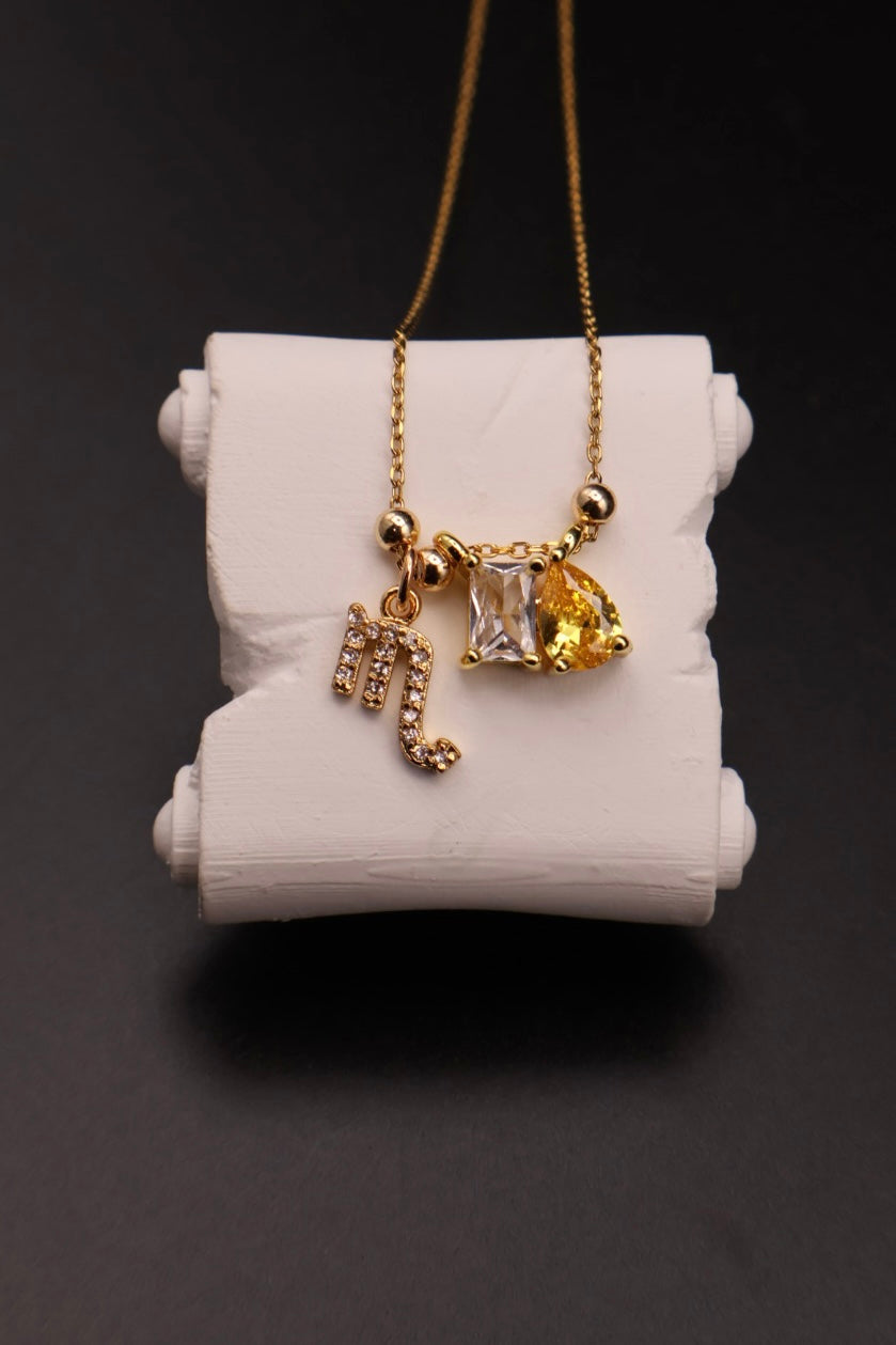A gold necklace with a letter 'M' initial pendant, a small rhinestone-embellished pendant, and a yellow heart-shaped crystal pendant, displayed on a white cylindrical stand against a dark background.