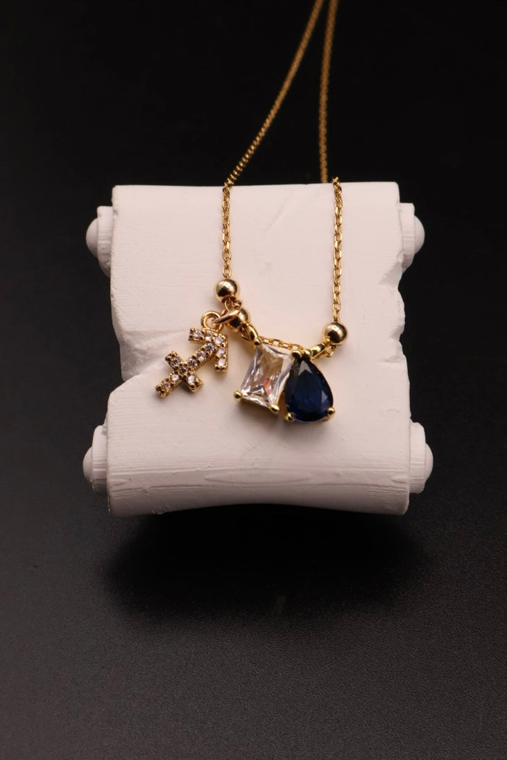 A gold chain necklace with a multi-charm pendant, displayed on a white cylindrical jewelry stand against a black background. The pendant features a small gold cross, a clear faceted gemstone, and a dark blue teardrop-shaped gemstone.