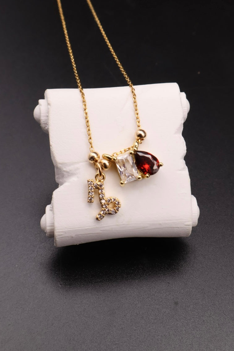 A gold necklace with a pendant featuring a clear rectangular gemstone, a dark red teardrop gemstone, and a small gold initial charm, displayed on a white cylindrical stand against a dark background.