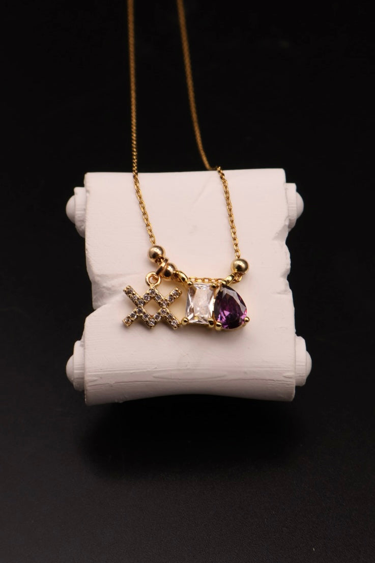A gold-tone necklace with a triple-charm pendant displayed on a white cylindrical stand against a black background. The pendant features a gold star, a clear faceted gemstone, and a teardrop-shaped purple gemstone.