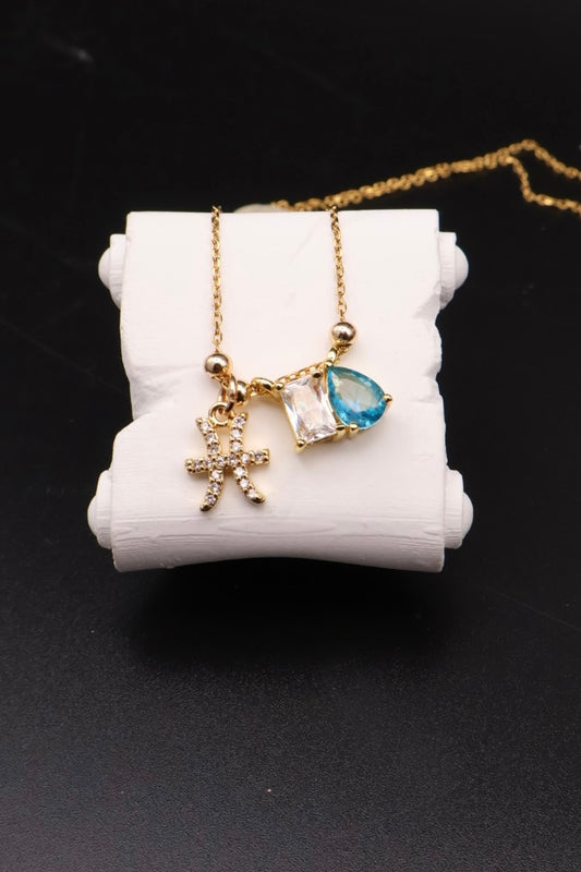 A gold chain necklace with a multi-charm pendant displayed on a white cylindrical stand against a black background. The pendant features a gold cross, a small clear faceted stone, and a teardrop-shaped blue gemstone.