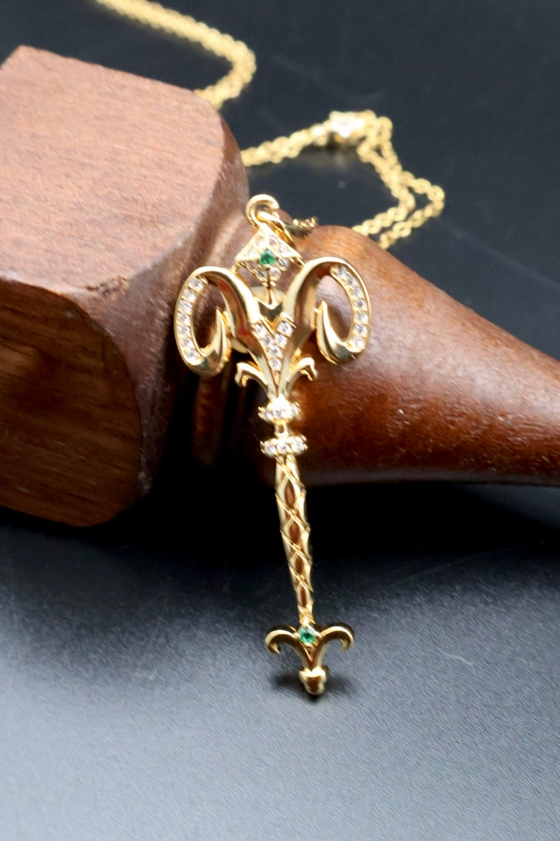 A gold pendant necklace with a decorative fleur-de-lis design, accented with small green gemstones and clear crystals, displayed on a dark surface next to a wooden cone.