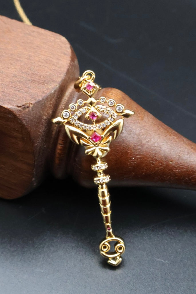 A gold-toned key-shaped pendant or charm with pink gemstone accents and small clear stones, displayed on a dark wooden cone against a dark background.