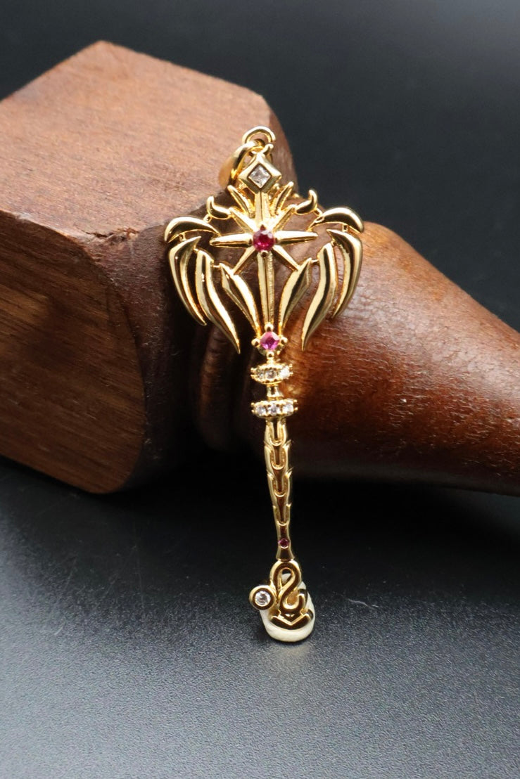 A gold pendant shaped like a decorative key or ornate tool, featuring small red gemstone accents, displayed on a dark wooden cone against a dark gray background.