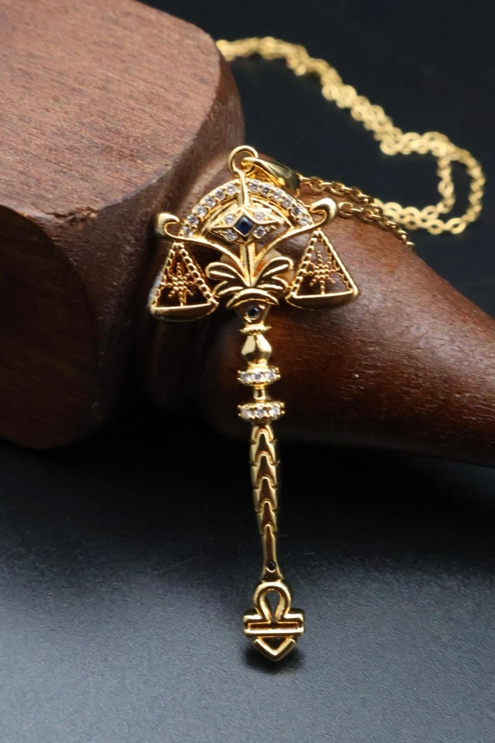 A gold-toned pendant necklace with a stylized animal head and dangling chain, displayed on a dark wooden stand against a dark background.