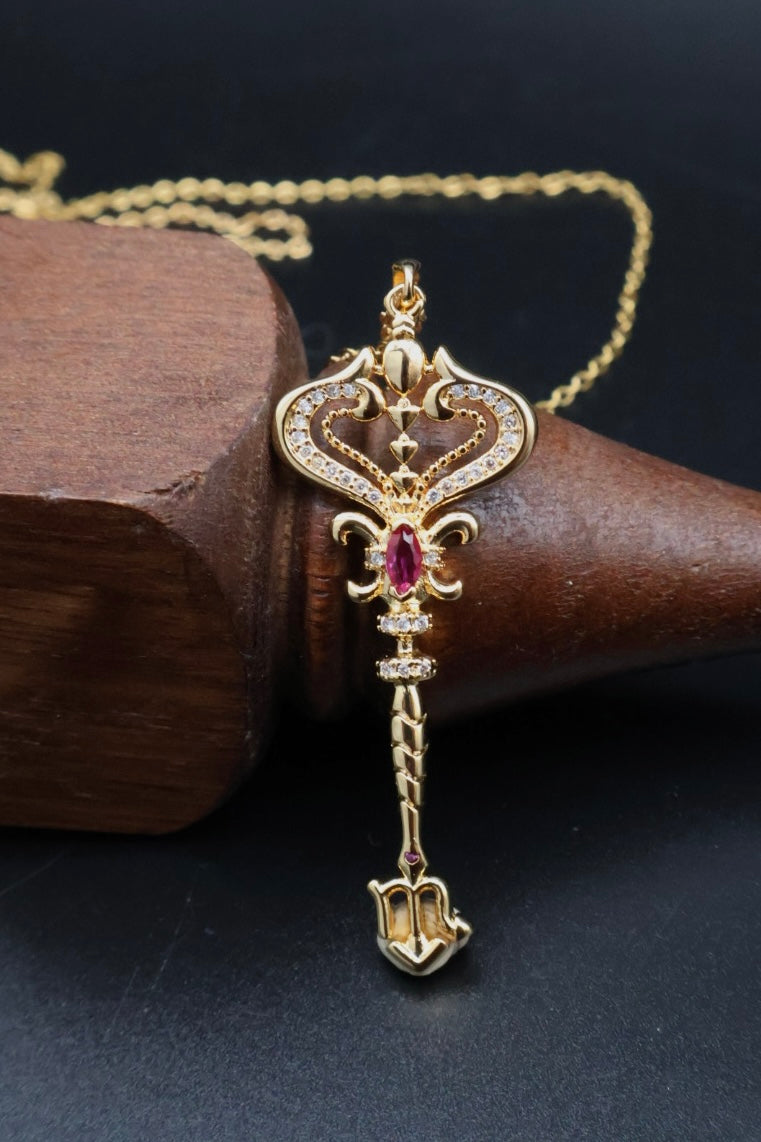 A gold pendant necklace with a decorative key-shaped design, accented with small clear stones and a central red gemstone, displayed on a dark wooden cone against a black background.