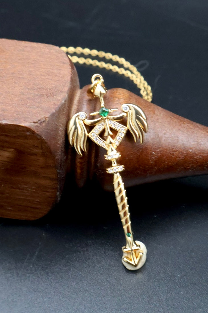 A gold-plated pendant necklace featuring a winged sword design with small green gemstone accents, resting on a dark wooden cone against a black background.