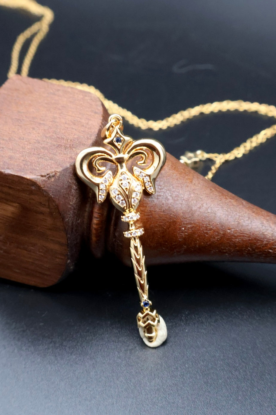 A gold-toned pendant necklace featuring a decorative key-shaped charm with a dangling teardrop charm, displayed on a dark wooden cone against a dark background.