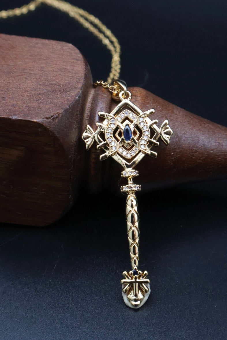 A gold pendant necklace with an ornate key-shaped design, featuring a small blue stone in the center and clear rhinestone accents, resting on a dark wooden block against a black background.