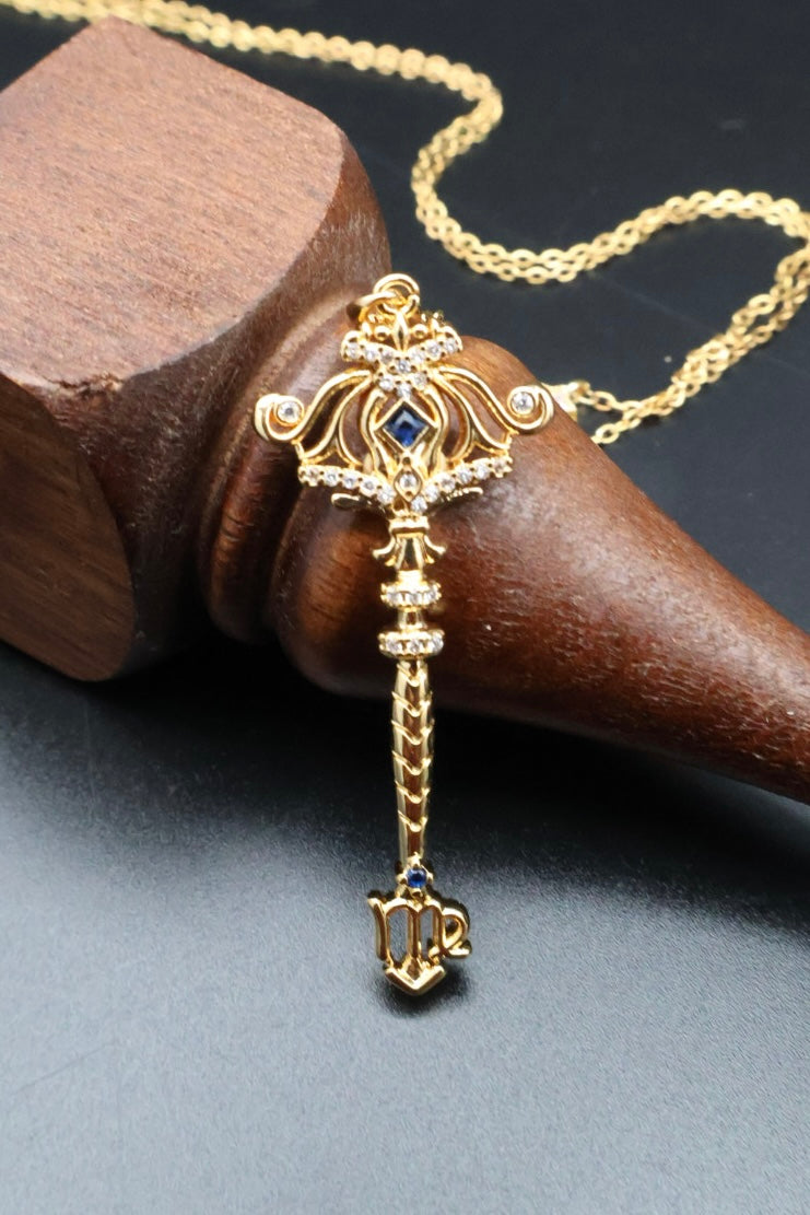 An ornate gold pendant necklace with a key-like design, small clear stones, and blue gemstone accents, displayed on a dark surface beside a wooden cone stand.