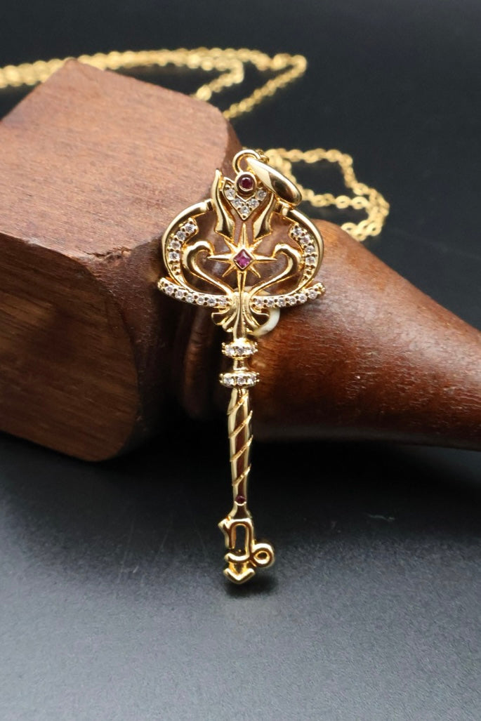 A gold key-shaped pendant necklace with a small red gemstone and clear rhinestones, displayed on a dark wooden cone against a dark gray background.