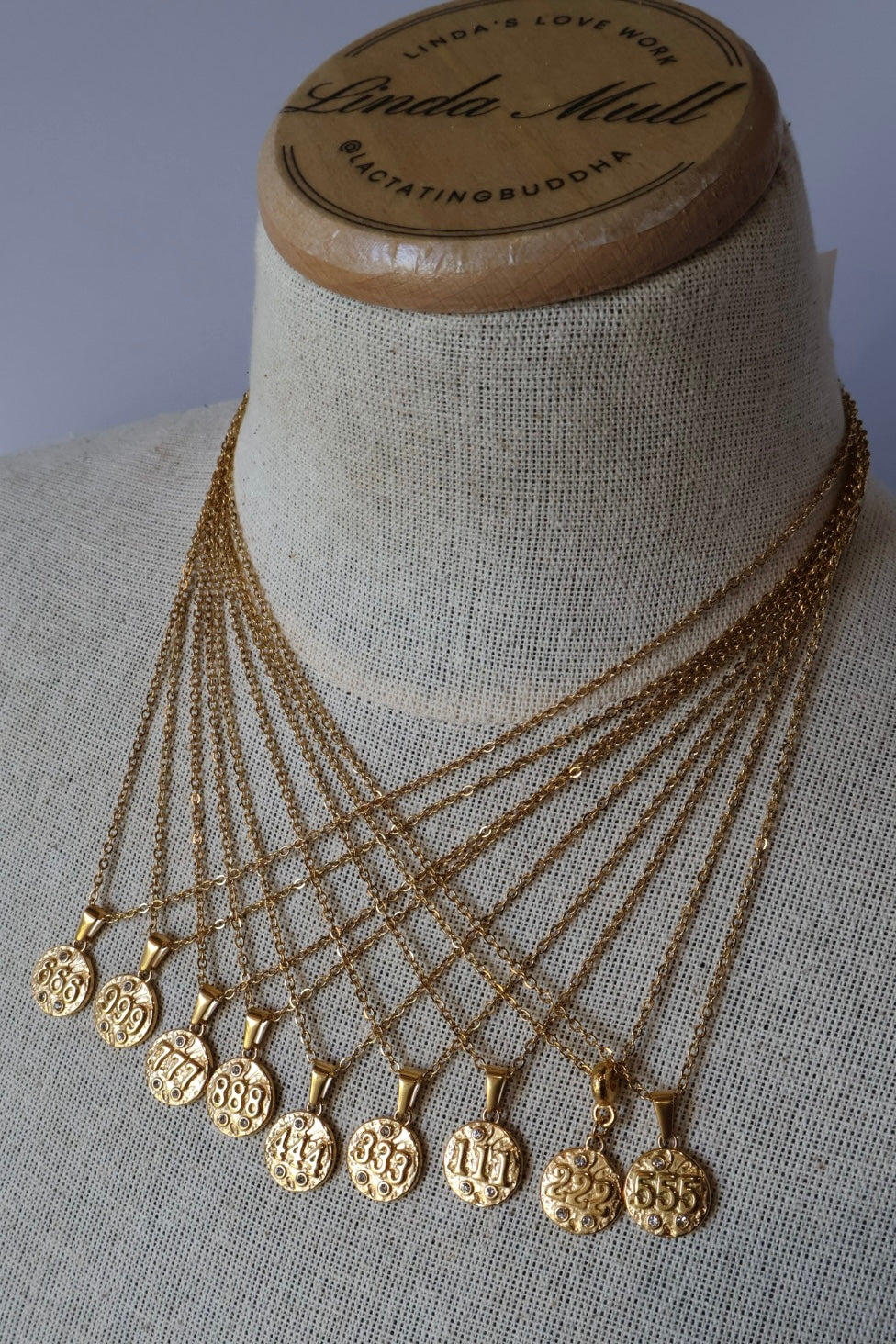 A set of gold-toned layered necklaces with engraved circular pendants displayed on a beige fabric-covered jewelry bust. The top of the bust shows a wooden label reading Linda Mull.