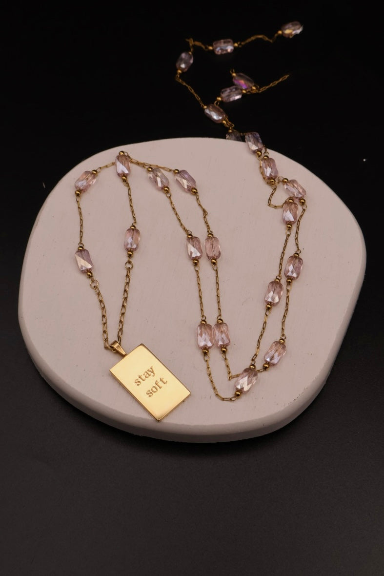 A gold chain necklace with pink faceted beads and a rectangular gold pendant engraved with 'stay soie', displayed on a round beige stone stand against a black background.