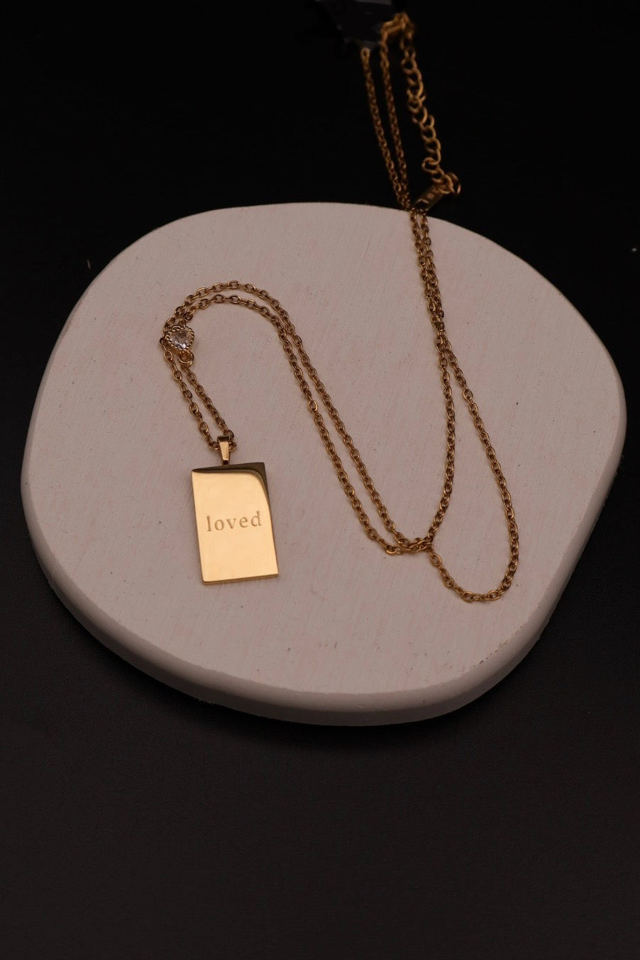 A gold-toned double-strand necklace with a rectangular pendant engraved with the word 'loved', displayed on a light gray stone tray against a black background.