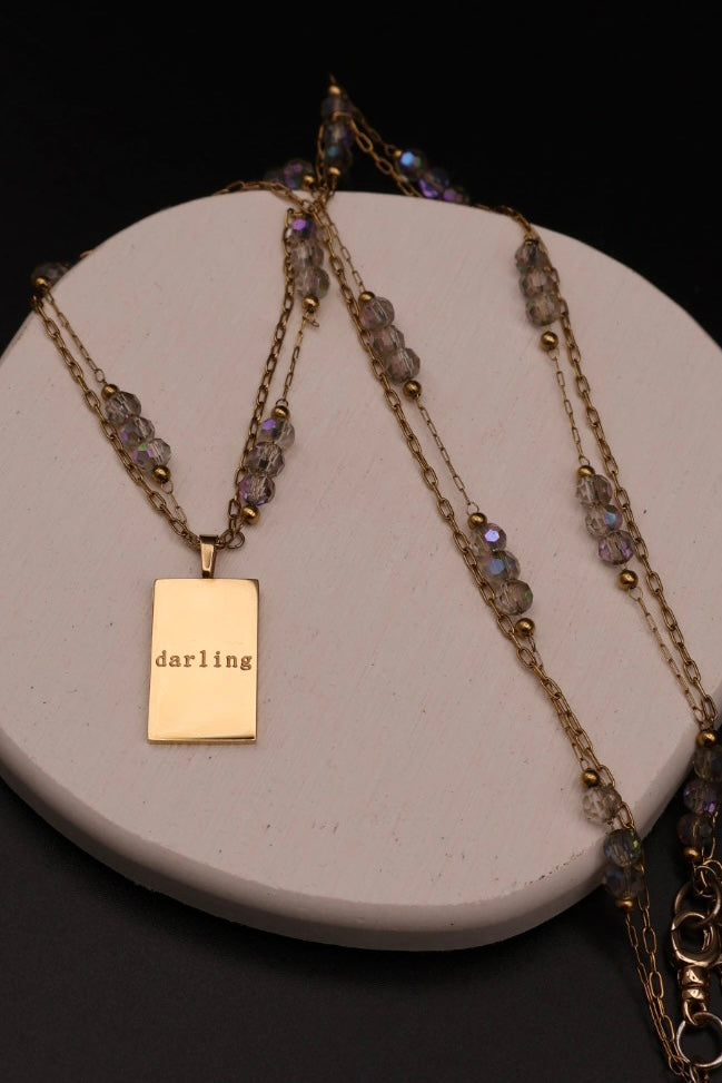 A gold-toned layered necklace with a rectangular pendant engraved with the word 'darling,' displayed on a round white stand against a black background.