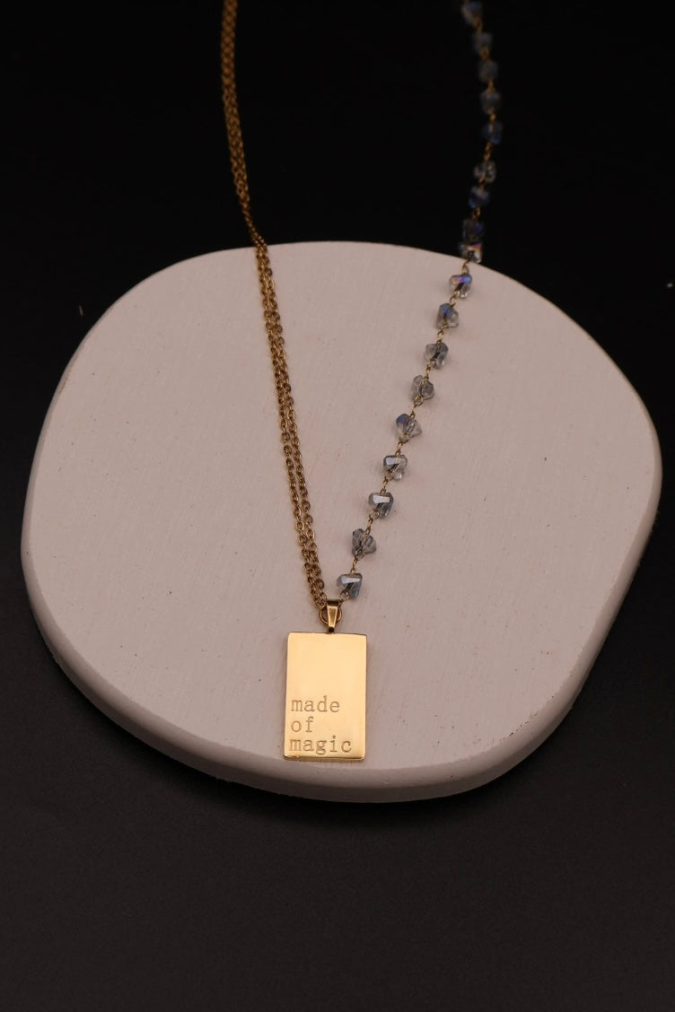 A gold-tone necklace with a rectangular engraved pendant reading 'made of magic' and a dangling strand of faceted gray-blue beads, displayed on a white ceramic stand against a black background.