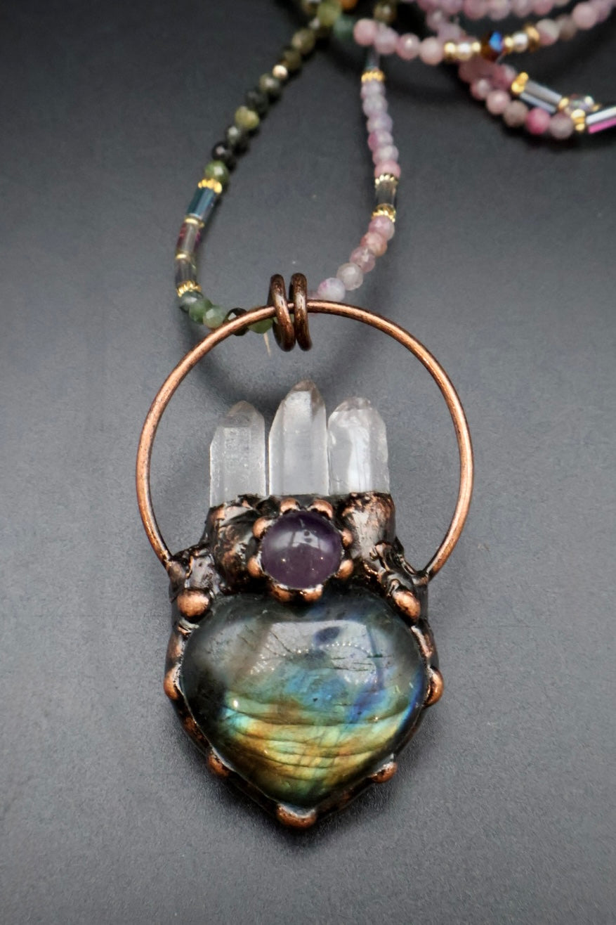 A beaded necklace with a large copper-toned hoop and a heart-shaped pendant featuring a iridescent labradorite stone, clear crystal points, and a purple amethyst accent, displayed on a dark gray background.