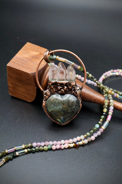 A heart-shaped labradorite pendant necklace with a copper ring and clear quartz points, displayed on a wooden stand against a dark background.