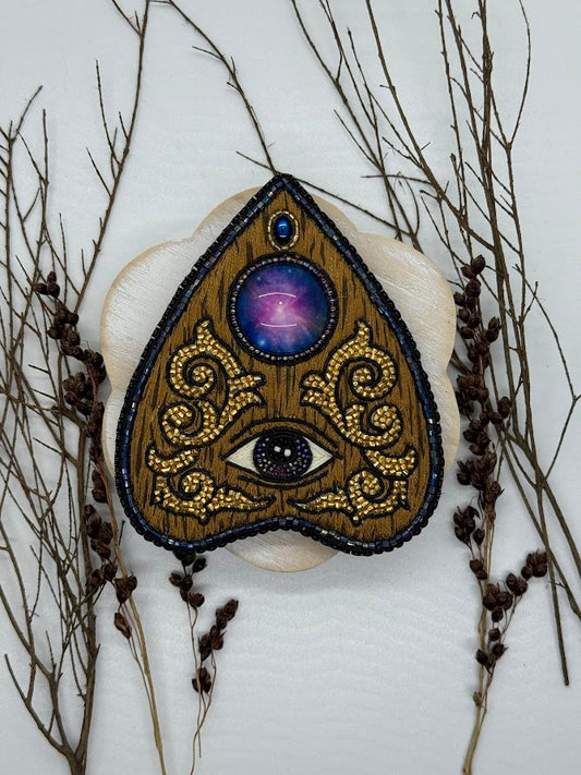 A picture of a planchette pin that looks like wood. the center stone looks like the galaxy. the center of the planchette shows an eye.