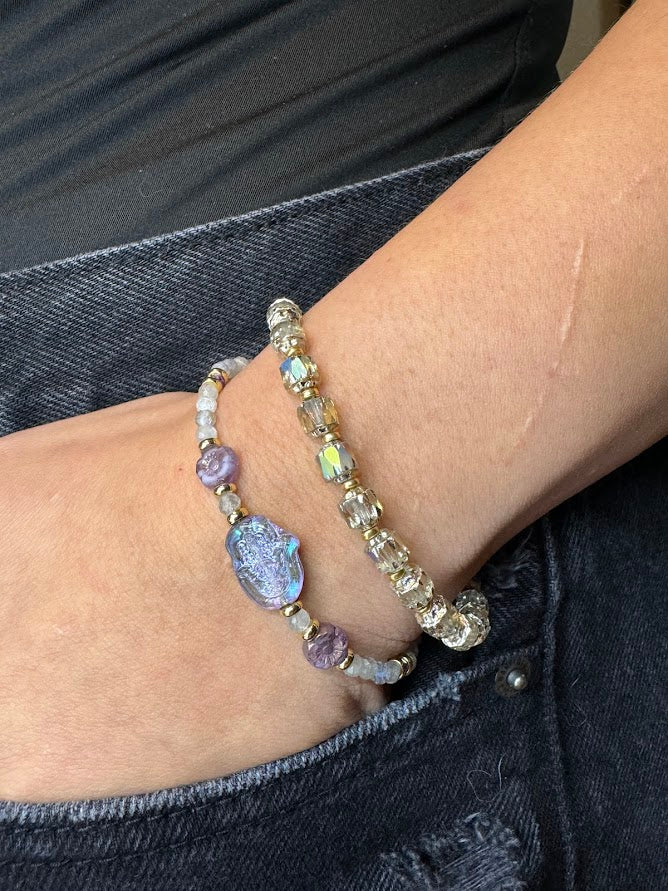 A wrist wearing a bracelet with labradorite beads, a hamsa hand focal, and two purple flower beads and a bracelet with baroque styled beads.