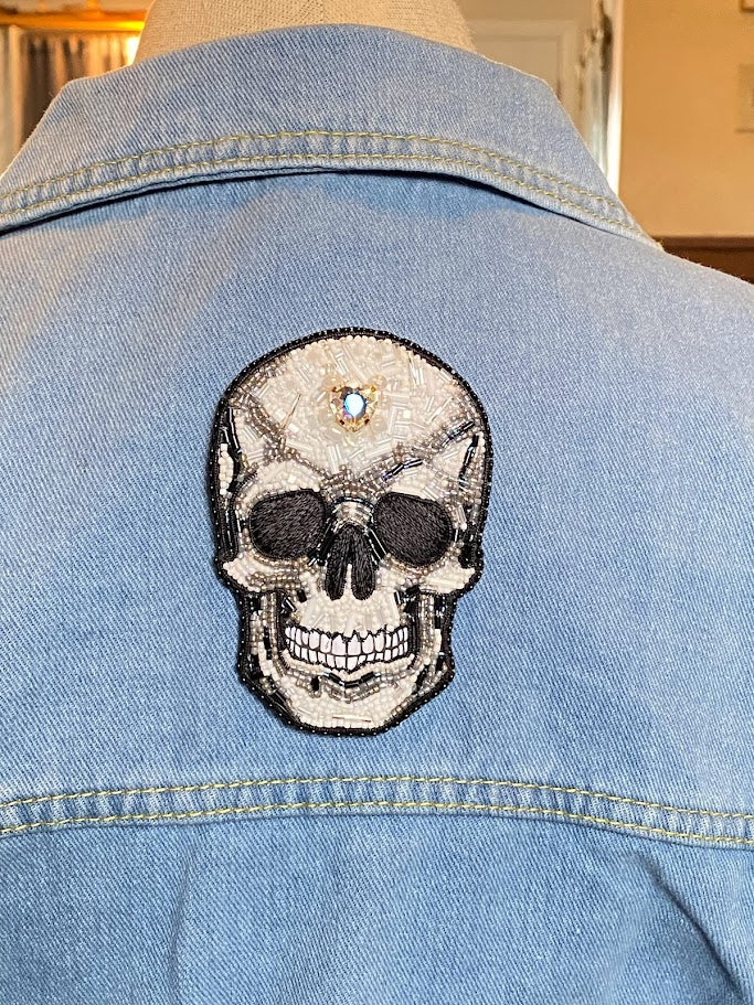 a skull pin on the back of a denim jacket. the pin is made with black, white, and gray beads. a heart shaped rhinestone is in the middle of the forehead.