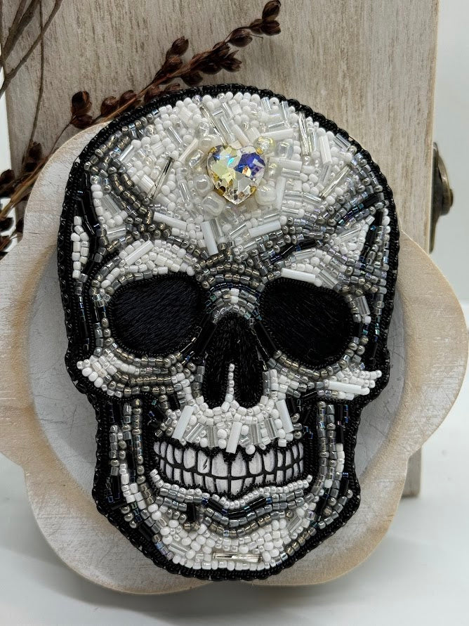 a close up of a skull pin made with black, white, and gray beads. a heart shaped rhinestone is in the middle of the forehead.