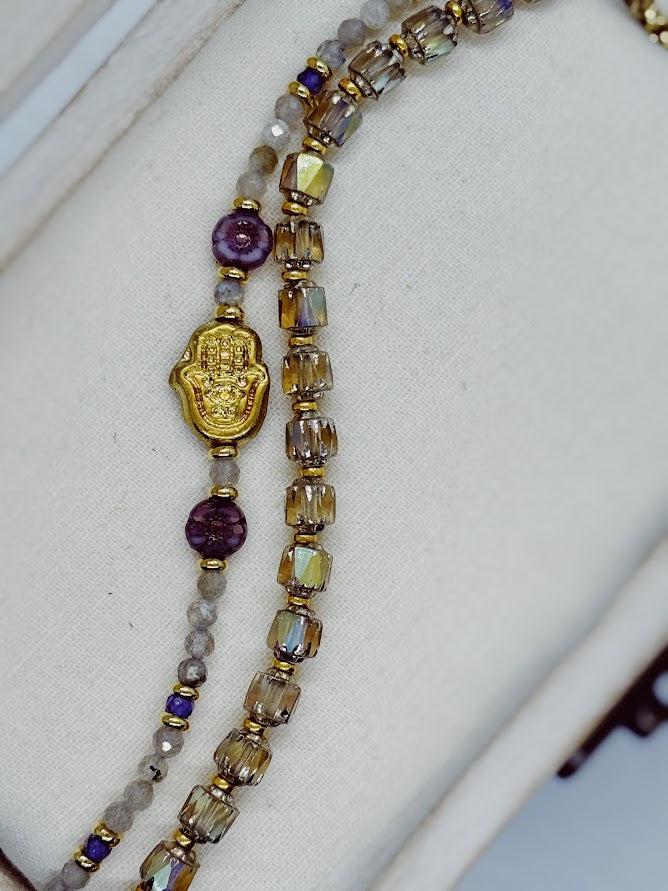 A closeup of a bracelet with labradorite beads, a hamsa hand focal, and two purple flower beads and a bracelet with baroque styled beads.