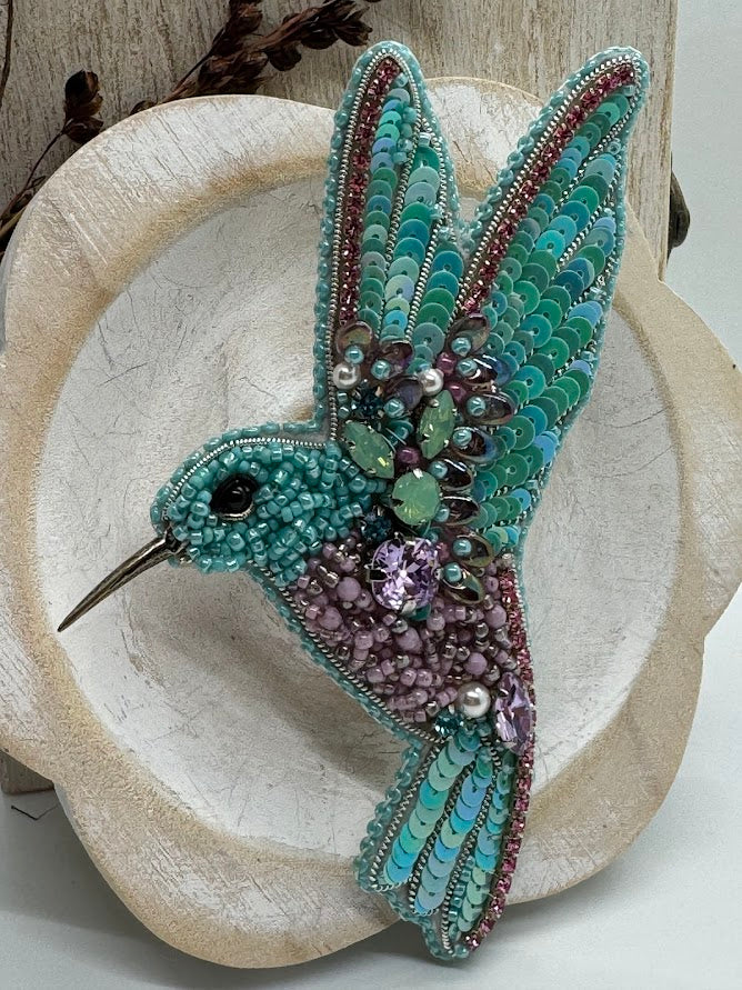 a closeup of A teal hummingbird pin made of beads and sequins.