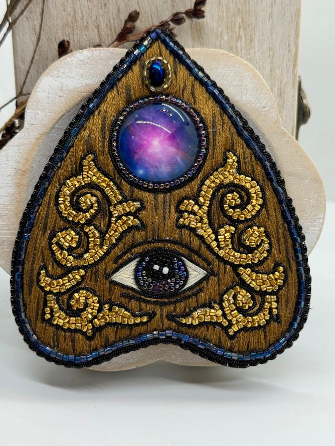 a closeup of planchette pin that looks like wood. the center stone looks like the galaxy. the center of the planchette shows an eye.