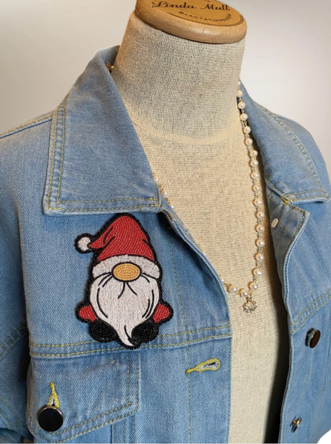 a bead embroidered pin of a Christmas gnome with a Santa hat on pinned to a denim jacket.