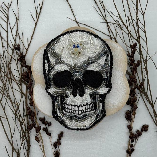 a skull pin made with black, white, and gray beads. a heart shaped rhinestone is in the middle of the forehead.