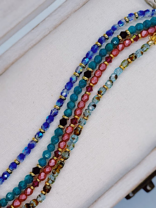 a bright blue bracelet with gold accents, a green bracelet with black bicone beads, a bright pink bracelet with pink bicone beads, a bracelet with brown and blue beads and gold accents.